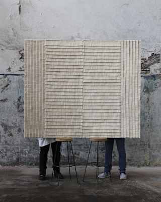 The legs of two men are visible behind a raised Estrella Wall Rug designed by Caralarga using repurposed raw cotton thread.