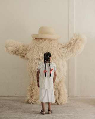 Seen from behind, a girl with long braids looks at a white fluffy monster designed by Fernando Laposse using agave fibres.