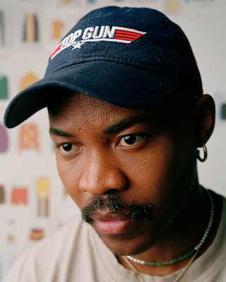 Close-up of designer Lukhanyo Mdingi, with a moustache and earring, wearing a black cap with the red and white Top Gun logo.