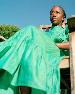 Low-angle image of designer Sindiso Khumalo sitting in a wooden garden chair wearing a high-necked pea-green dress.