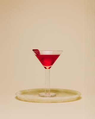 Eye-level view of a glowing, deep red Rose Martini cocktail, garnished with a petal, served in a thick-stemmed Martini glass.