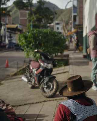 A man in a fedora checks his phone by a shop opening, watched by a local woman wearing a brown bolero hat, seen from behind.