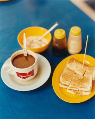 A plate of Kaya Toast sliced into six pieces with a sandwich pick seen from above on blue cloth and a cup of hot chocolate.