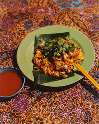 A crispy oyster omelette is served on a green plate, set on a Malay fabric cloth with chopsticks and dipping sauce.