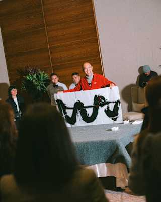 Artist Wu Jian'an, in glasses and red top, holds up one of his ink artworks, in a view from the audience between two heads.