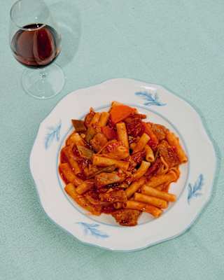 Looking down on a plate of rigatoni pasta in a rich vegetable ragout, served with a glass of red wine on a pale blue tablecloth.