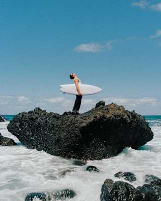 A man with a shortie surfboard under his arm stands, looking at the sky ,on a large basaltic rock surrounded by foaming sea.
