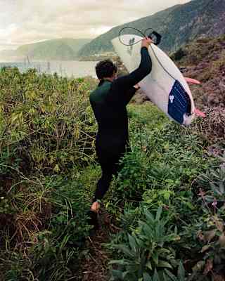 A man in a wetsuit carries his surfboard, with pink fins, through Madeira's undergrowth towards the ocean, seen from behind.