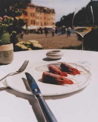 A knife and fork rest against a plate with three Santa Margherita prawns at a table with white wine set back from the marina.