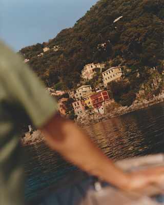 Angled image of Portofino's red, pink and yellow marina-side buildings, seen over the arm of a man on a boat in soft-focus.