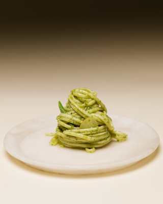A perfect twirled mound of trenette pasta in a fresh, vibrant pesto sauce, served on a simple white plate, seen in close-up.