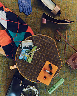 looking down on a round Louis Vuitton bag scattered with items including clothes, camera, lipstick, postcard, hipflask and comb.