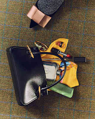 Looking down on a black handbag which spills its contents, comprising a hip flask, green comb, yellow scarf and red lipstick.