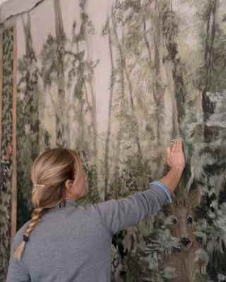 Elena Carozzi, with blond hair in a plait, rests her hand on a large hanging paper with botanical motifs, seen from the side.