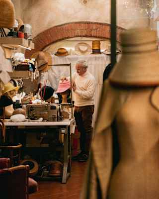 Hatmaker Antonio Gatto places a pink hat on a model head in a studio filled with his creations and tools, seen past a dummy.