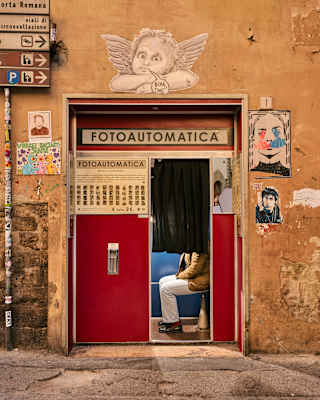 A person, face obscured by a curtain, sits in a restored 1960s Fotoautomatica photo booth set into a street wall in Florence.