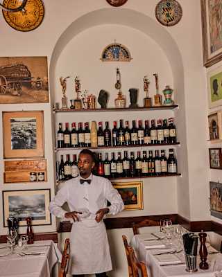 A smart waiter in whites polishes a glass in Trattoria Cammillo, surrounded by historic pictures and shelves of wine bottles.