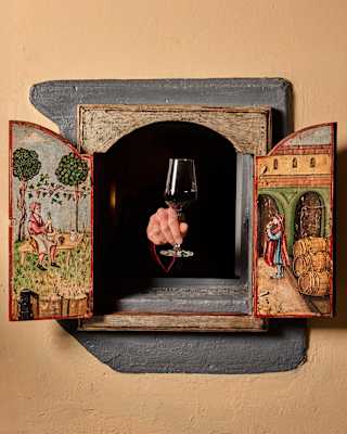A wine window wall hatch, doors open and their insides painted with rustic scenes, reveals a hand holding glass of red wine.