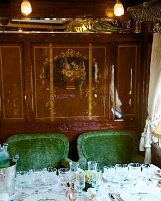 Two plush green chairs sit beneath a floral marquetry panel, seen over a table filled with glasses in the Etoile du Nord car.