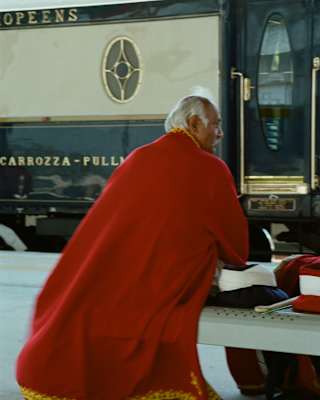 A grey-haired man sits on a platform bench in front of the train, in a long red cloak with gold piping, seen from behind.