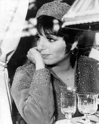 A woman in a beaded outfit sits at a table with glassware, resting her chin on her hand and looking to the side. Draped curtains and a lampshade are visible in the background.