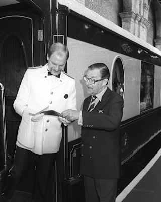 A man in a white uniform stands at a train door, holding a document, whilst another man in a dark suit points at the paper. They appear to be discussing something on a railway platform.