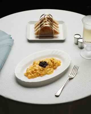 A white oval plate with creamy scrambled eggs topped with caviar sits on a round table beside a fork, a glass of drink, a folded blue serviette, and a plate of sliced bread.