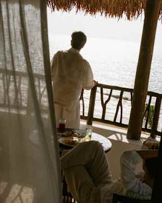 High angle view of a man in white top and trousers leaning on his balcony rail as he gazes at sun-bleached sea below.