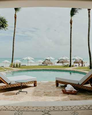 Four loungers surround a private pool set back from the sand, sea and beach parasols, seen from Villa Maroma's colonnade.