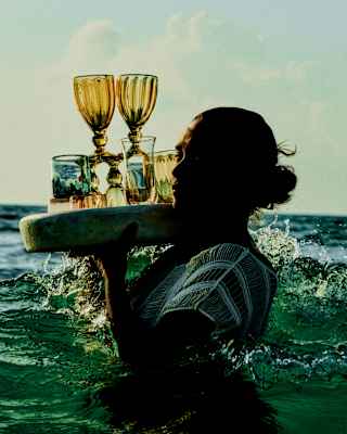A woman carries a round tray of drinks and unusual glasses on her right shoulder as she wades through waist-high ocean water.