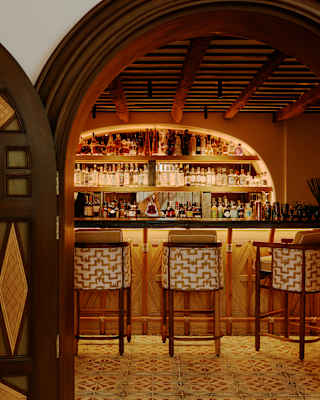 View through Bambuco's arched doorway of three wicker stools at the sub-lit counter of the bar, with backlit bottle shelves.