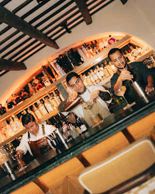 Angled shot of three male bartenders preparing cocktails at Bar Bambuco, illuminated by the lit bottle shelves behind.
