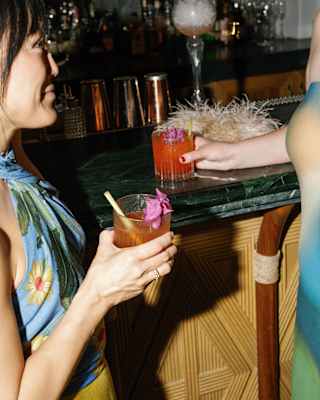A woman in a floral top and yellow skirt stands at a bar with a flower-garnished drink as she chats with a friend off-camera.