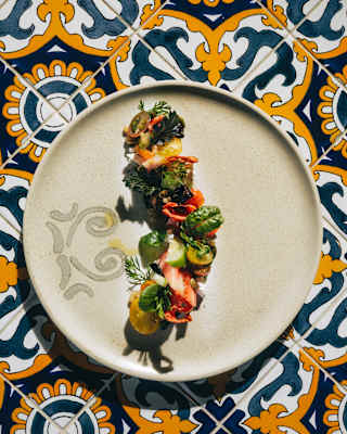A vibrant starter of tomatoes and herbs arranged on a plate in a line, seen from above on an orange and blue-tiled surface.