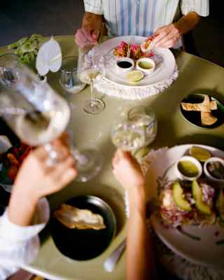 A hand holds a glass of white wine at a table of diners enjoying fresh tacos with vibrant toppings, guacamole and bean dip.