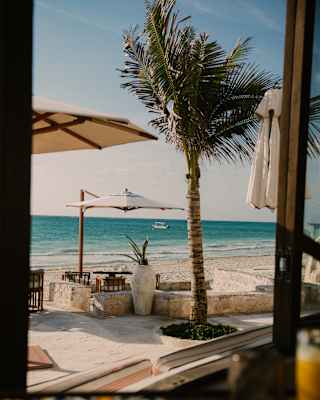 A cantilever parasol shades an outside, waterfront table set on the white sand beach, viewed through a window at Casa Mayor.