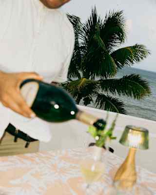 Angled image of a waiter pouring wine at a table in soft-focus foreground, with crisp views of a palm tree and ocean behind.