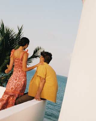A woman in red dress stands by a man in a yellow shirt, sitting on the edge of the roof terrace overlooking the rolling sea.
