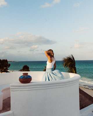 A woman in a white top and blue skirt stands at Maroma's El Mirador circular white-walled viewpoint, gazing at the azure sea.