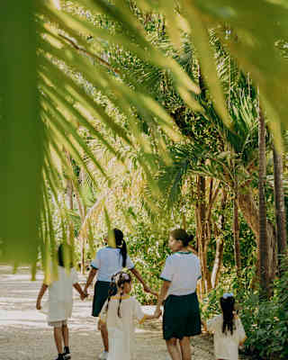 Two kids club staff members hold the hands of children as they wander along a garden path, seen from behind a palm frond.