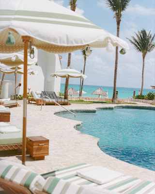The pool's water complements the electric blue of the sea beyond, divided by palms and parasols, seen from behind a lounger.