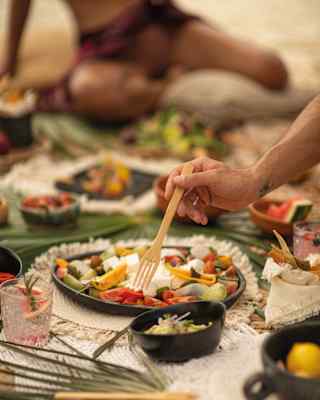 A person uses chopsticks to pick food from a platter during an outdoor meal on sand, with tropical leaves and various dishes spread about. Another person is seated in the background.
