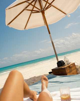 View of the white beach and azure sea, taken from the waist of a woman whose legs stretch out on towels beneath a parasol.