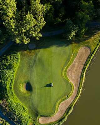 Aerial view of a buggy on a flat mown area of an emerald-green golf course with a wavy sand bunker bordering a lake.