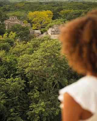 High-angle view of the forests and stone monuments of the Ek Balam ruins, seen past a woman in soft-focus.