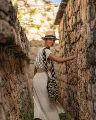 A woman in a straw hat and white dress glances back at the camera as she walks through a narrow lane in the Ek Balam Ruins.