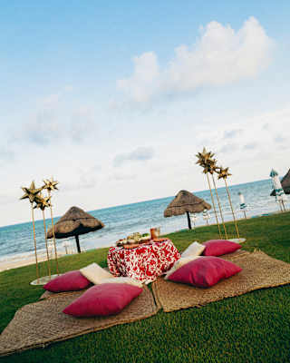 A seaside picnic set-up with red cushions, a patterned blanket, and a low table on grass. Two thatched parasols and decorative windmills stand nearby with the sea and blue sky in the background.