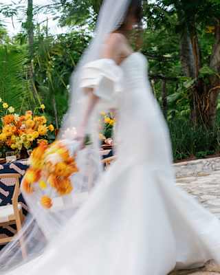 A bride in a long dress and veil creates a beautiful wedding-white blur as she walks with a bouquet of bright orange flowers.
