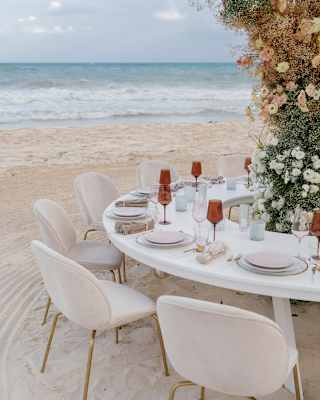 View over the beach, past a doughnut-shaped party table with red glasses and plates, and six chairs circling a floral column.