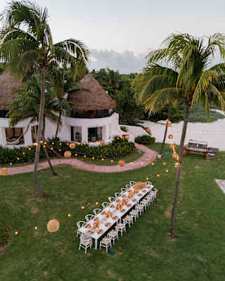 Seen at dusk, light strings hang between palms on a lawn above a long celebration table for twenty guests, seen from above.